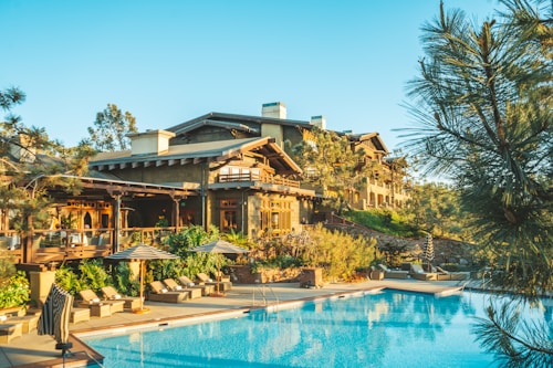 A luxurious resort with wooden architecture surrounded by lush greenery is visible. The main building has a rustic design with multiple levels and balconies. In front of the building, there is a large, inviting swimming pool with lounge chairs and umbrellas arranged neatly beside it. Tall trees and shrubs enhance the natural look of the area, and the sky is clear and blue.