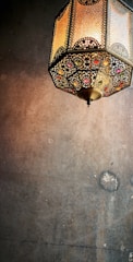 A decorative hanging lantern with intricate metalwork and colorful glass embellishments casts a warm glow against a textured wall.