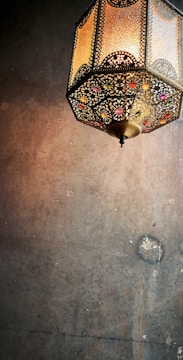 A decorative hanging lantern with intricate metalwork and colorful glass embellishments casts a warm glow against a textured wall.