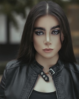 A woman with striking makeup and bold eyebrows is wearing a black leather jacket. Her long, dark hair frames her face, and she gazes directly at the camera with an intense expression. The background is softly blurred, drawing attention to her features.