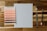 A wooden surface displays a Pantone color specifier with various orange and pink hues. Beside it lies a blank sheet of paper, a small Pantone color swatch, a metal ruler, and a pencil.
