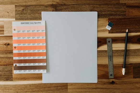 A wooden surface displays a Pantone color specifier with various orange and pink hues. Beside it lies a blank sheet of paper, a small Pantone color swatch, a metal ruler, and a pencil.