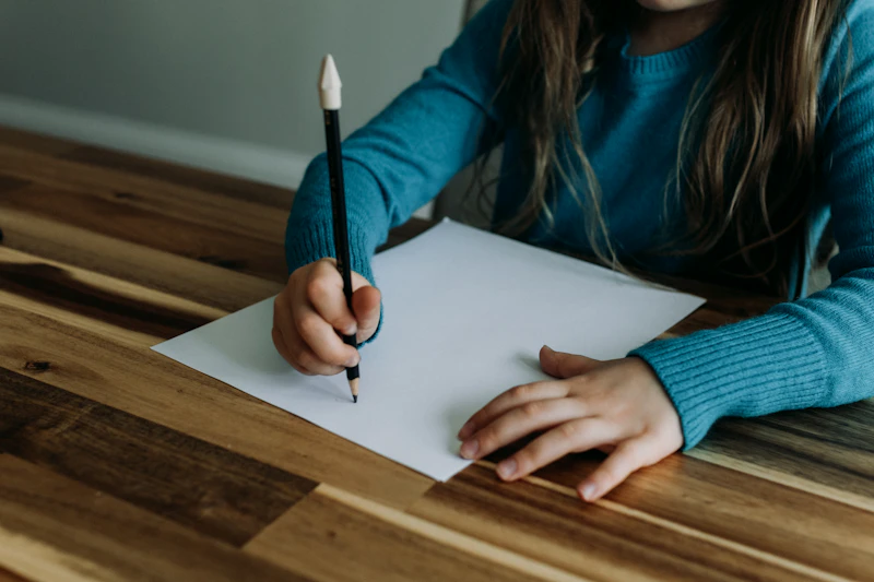 A girl's hands drawing with a pencil on white paper at a wooden table
