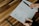 A person is sketching a wireframe layout on a piece of paper, using a ruler for precise lines. The sheet has designs for a user interface layout, including menu and content sections. The person is holding a pen and wearing a ring, working on a wooden table with a modern design.