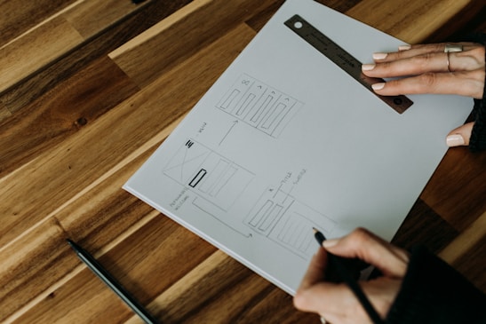 A person is sketching a wireframe layout on a piece of paper, using a ruler for precise lines. The sheet has designs for a user interface layout, including menu and content sections. The person is holding a pen and wearing a ring, working on a wooden table with a modern design.