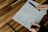 A person is sketching a wireframe layout on a piece of paper, using a ruler for precise lines. The sheet has designs for a user interface layout, including menu and content sections. The person is holding a pen and wearing a ring, working on a wooden table with a modern design.