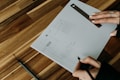 A person is sketching a wireframe layout on a piece of paper, using a ruler for precise lines. The sheet has designs for a user interface layout, including menu and content sections. The person is holding a pen and wearing a ring, working on a wooden table with a modern design.
