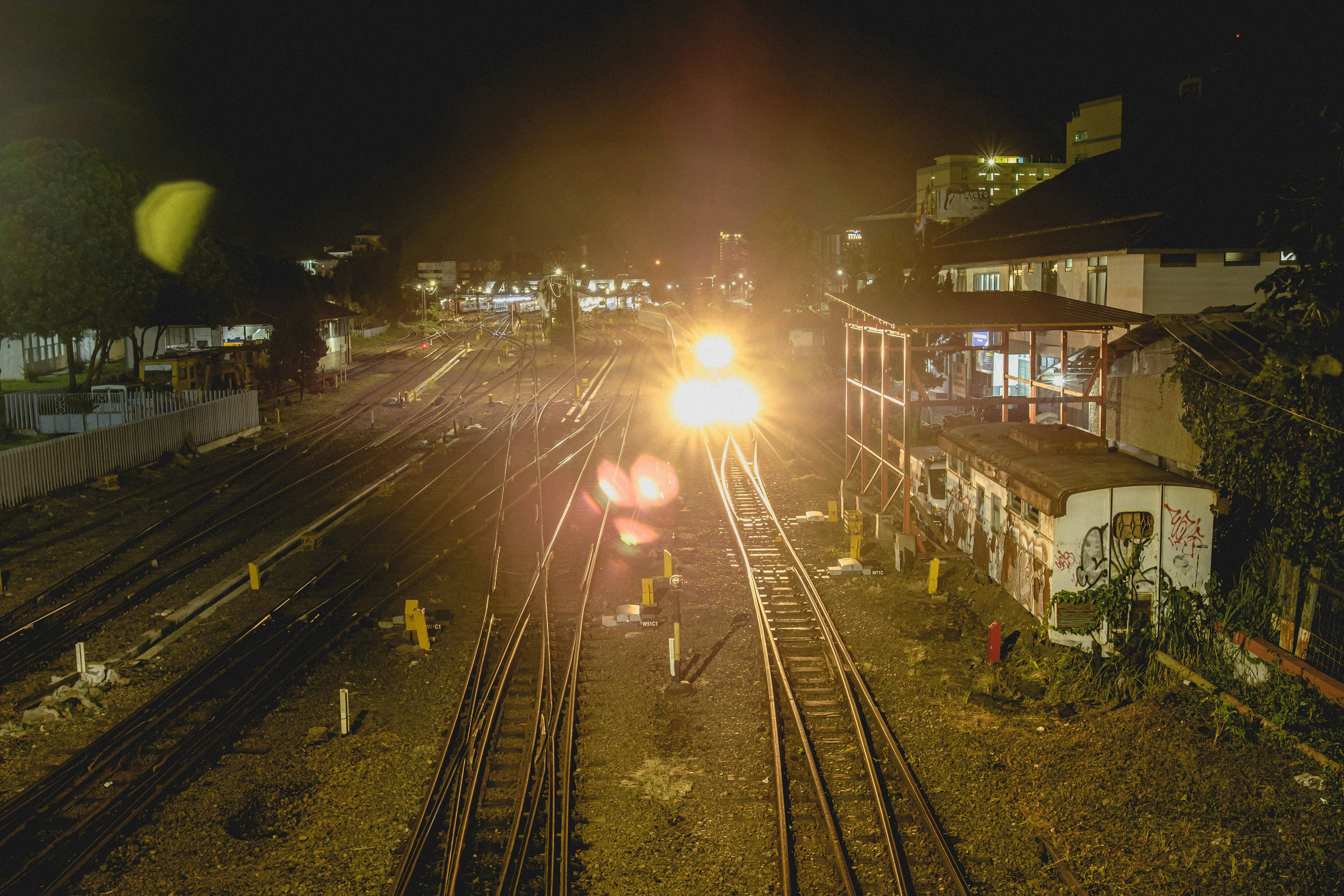 Farol de um trem se aproximando à noite pelos trilhos, com casas iluminadas ao lado da linha