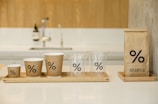 A set of cups and a coffee bag are arranged on wooden trays. Each item features a minimalist design with a percentage symbol. The cups vary in size and include both paper and plastic materials. A modern kitchen faucet and soap dispenser are blurred in the background.