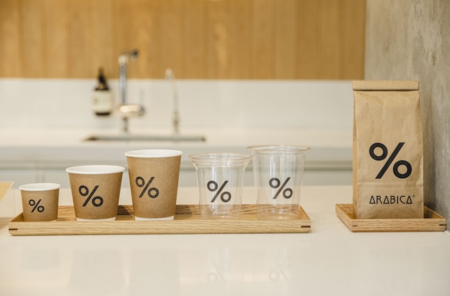 A set of cups and a coffee bag are arranged on wooden trays. Each item features a minimalist design with a percentage symbol. The cups vary in size and include both paper and plastic materials. A modern kitchen faucet and soap dispenser are blurred in the background.