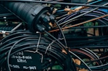 A tangled cluster of various cables and wires, some labeled with text. The cables are wrapped around and intertwined with a cylindrical connector device, with parts that are metallic and rubber-coated. The background is filled with more wires and some blurred elements of the environment, possibly outdoor.