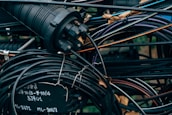 A tangled cluster of various cables and wires, some labeled with text. The cables are wrapped around and intertwined with a cylindrical connector device, with parts that are metallic and rubber-coated. The background is filled with more wires and some blurred elements of the environment, possibly outdoor.