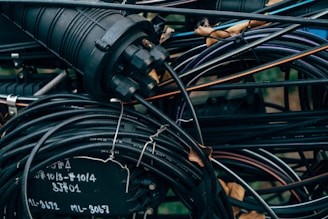 A tangled cluster of various cables and wires, some labeled with text. The cables are wrapped around and intertwined with a cylindrical connector device, with parts that are metallic and rubber-coated. The background is filled with more wires and some blurred elements of the environment, possibly outdoor.