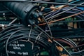 A tangled cluster of various cables and wires, some labeled with text. The cables are wrapped around and intertwined with a cylindrical connector device, with parts that are metallic and rubber-coated. The background is filled with more wires and some blurred elements of the environment, possibly outdoor.