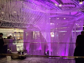 Intricate crystal bead curtains hang lavishly with a purple and pink lighting scheme, providing a luxurious and elegant atmosphere. The glass table and chair add to the sophisticated ambiance, while silhouettes of people are visible, suggesting patrons enjoying their surroundings.