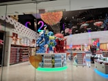 A brightly lit candy store showcasing a large, colorful candy-themed sculpture at its entrance. The interior features vibrant decorations, including walls lined with various candy products and eye-catching displays. The ceiling is adorned with a rainbow-like installation, adding to the playful and whimsical atmosphere.