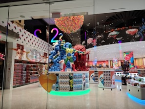 A brightly lit candy store showcasing a large, colorful candy-themed sculpture at its entrance. The interior features vibrant decorations, including walls lined with various candy products and eye-catching displays. The ceiling is adorned with a rainbow-like installation, adding to the playful and whimsical atmosphere.