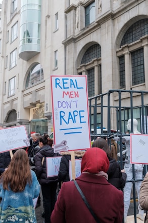 A group of people is gathered for a protest in an urban setting. They are holding signs with messages advocating against rape and promoting women's rights. The background features a building with classical architecture.