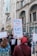 A group of people is gathered for a protest in an urban setting. They are holding signs with messages advocating against rape and promoting women's rights. The background features a building with classical architecture.