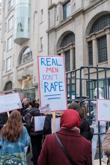 A group of people is gathered for a protest in an urban setting. They are holding signs with messages advocating against rape and promoting women's rights. The background features a building with classical architecture.