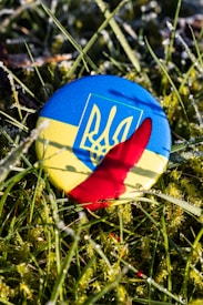 A round badge displaying the Ukrainian flag and coat of arms lies on grass, partially covered by a splash of red liquid. The grass appears frosty or dewy, creating a textured background with small droplets.