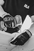 A black and white image captures a person wearing gloves using a power drill. The focus is on the hands and the drill, suggesting a construction or woodworking activity. The person is wearing a shirt with a logo on it.