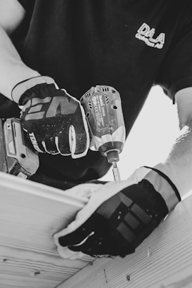 A black and white image captures a person wearing gloves using a power drill. The focus is on the hands and the drill, suggesting a construction or woodworking activity. The person is wearing a shirt with a logo on it.