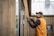 A man wearing an orange vest and black cap uses a power drill to work on a wooden wall of a building. He is focused on his task, and the background shows a metal roof and windows.