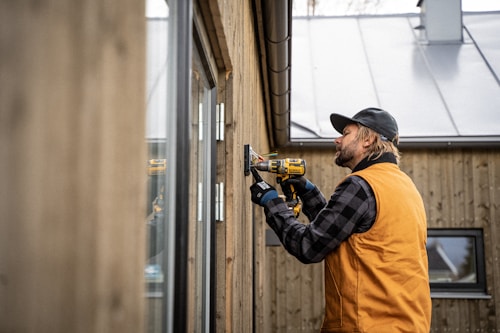 A man wearing an orange vest and black cap uses a power drill to work on a wooden wall of a building. He is focused on his task, and the background shows a metal roof and windows.