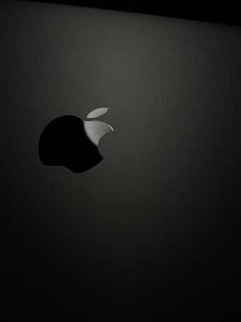 A sleek, minimalist design featuring a prominent, glossy apple logo against a matte dark background. The logo has a distinctive bite mark and is illuminated softly, creating a subtle reflection.