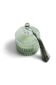 A glass jar with ribbed detailing containing a light green substance, topped with a matching lid featuring a metallic spherical handle. Attached to the handle is a decorative green tassel.