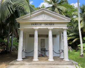 A small, white family mausoleum with four classical columns and a triangular pediment featuring the name 'Famille Dauban'. It is surrounded by lush tropical vegetation, including tall palm trees. The entrance is chained off, with some inscriptions visible on the walls inside.