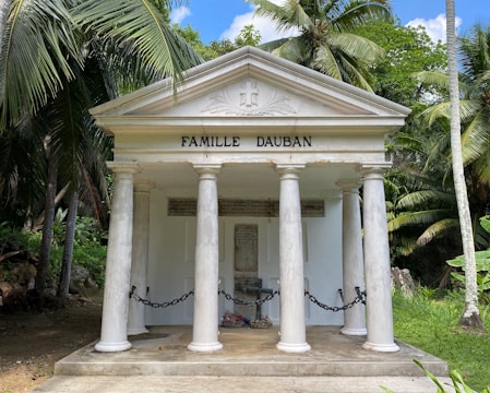 A small, white family mausoleum with four classical columns and a triangular pediment featuring the name 'Famille Dauban'. It is surrounded by lush tropical vegetation, including tall palm trees. The entrance is chained off, with some inscriptions visible on the walls inside.