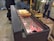 The image features a traditional setup for baking chimney cakes on a rotating spit over glowing charcoal embers. Three dough cakes with varying degrees of browning are placed on the spit. Nearby, individuals stand watching the process, and there are wooden handles and other cooking tools in the background.