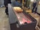 The image features a traditional setup for baking chimney cakes on a rotating spit over glowing charcoal embers. Three dough cakes with varying degrees of browning are placed on the spit. Nearby, individuals stand watching the process, and there are wooden handles and other cooking tools in the background.