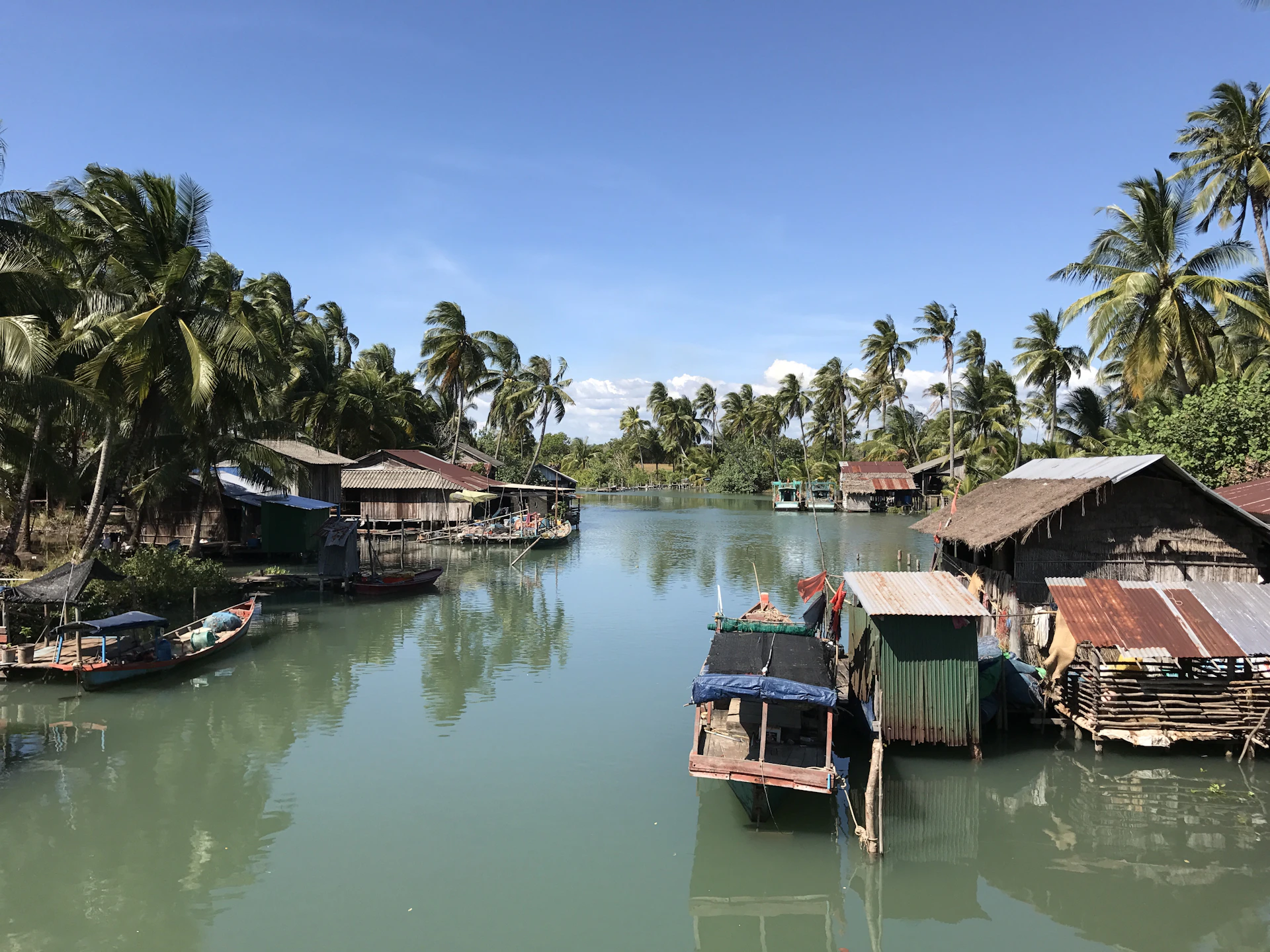 A serene riverside village scene showing Myènè houses on stilts with lush greenery and calm water reflections.