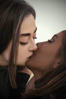 Two people share an intimate kiss, with their faces close together and eyes gently closed. The background is softly blurred, highlighting their connection.