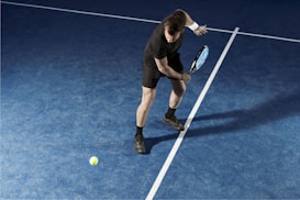 A tennis player in action, dressed in black sportswear, is captured mid-swing on a blue tennis court. The court is crossed by white lines and the player's shadow is visible on the court. A tennis ball is in motion near the player.