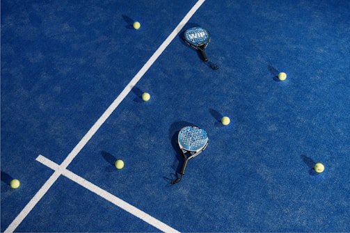 A blue court features white boundary lines with two padel rackets and several yellow tennis balls scattered across the surface.
