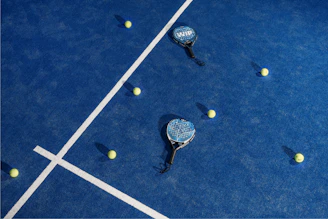 A selection of padel balls in a transparent container with a court and players in soft focus behind.