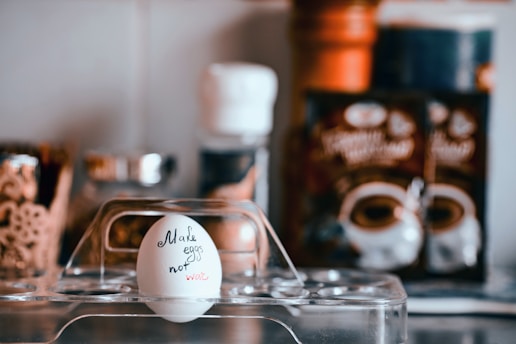 An egg is placed in a clear egg holder, with the words 'Make eggs not war' written on it. The background contains blurred kitchen items like spice jars and food packages, creating a domestic kitchen setting.