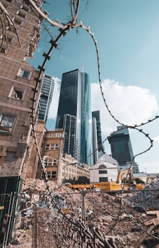 An urban scene featuring a construction or demolition site, with rubble and debris in the foreground. Skyscrapers tower in the background, framed by a barbed wire fence running diagonally across the image. The scene includes a mix of modern and older buildings, highlighting a contrast between development and decay.