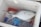 A fish is placed inside a cooler box alongside a sealed clear plastic bottle with a blue cap. The cooler box is white and has some red stains on its sides. The fish appears to have a few black spots on its body.