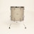 A standing drum with a textured beige surface is centered against a plain light background. It features chrome hardware including lugs, rims, and legs that extend to the floor to support the drum. The drum has a minimalist and vintage appearance.