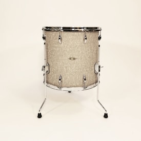 A standing drum with a textured beige surface is centered against a plain light background. It features chrome hardware including lugs, rims, and legs that extend to the floor to support the drum. The drum has a minimalist and vintage appearance.