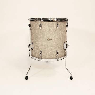 A standing drum with a textured beige surface is centered against a plain light background. It features chrome hardware including lugs, rims, and legs that extend to the floor to support the drum. The drum has a minimalist and vintage appearance.