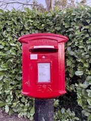 A red Royal Mail postbox stands prominently against a background of dense, green foliage. The postbox features a slot for letters and displays collection times on its front panel, along with the distinctive ER insignia.