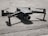 A small gray quadcopter drone with labeled 'Mavic 2' rests on a rough concrete surface. The drone has four rotors, with two angled slightly upward. Its compact form and camera at the front are highlighted by the shadows cast beneath it.