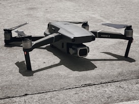 A small gray quadcopter drone with labeled 'Mavic 2' rests on a rough concrete surface. The drone has four rotors, with two angled slightly upward. Its compact form and camera at the front are highlighted by the shadows cast beneath it.