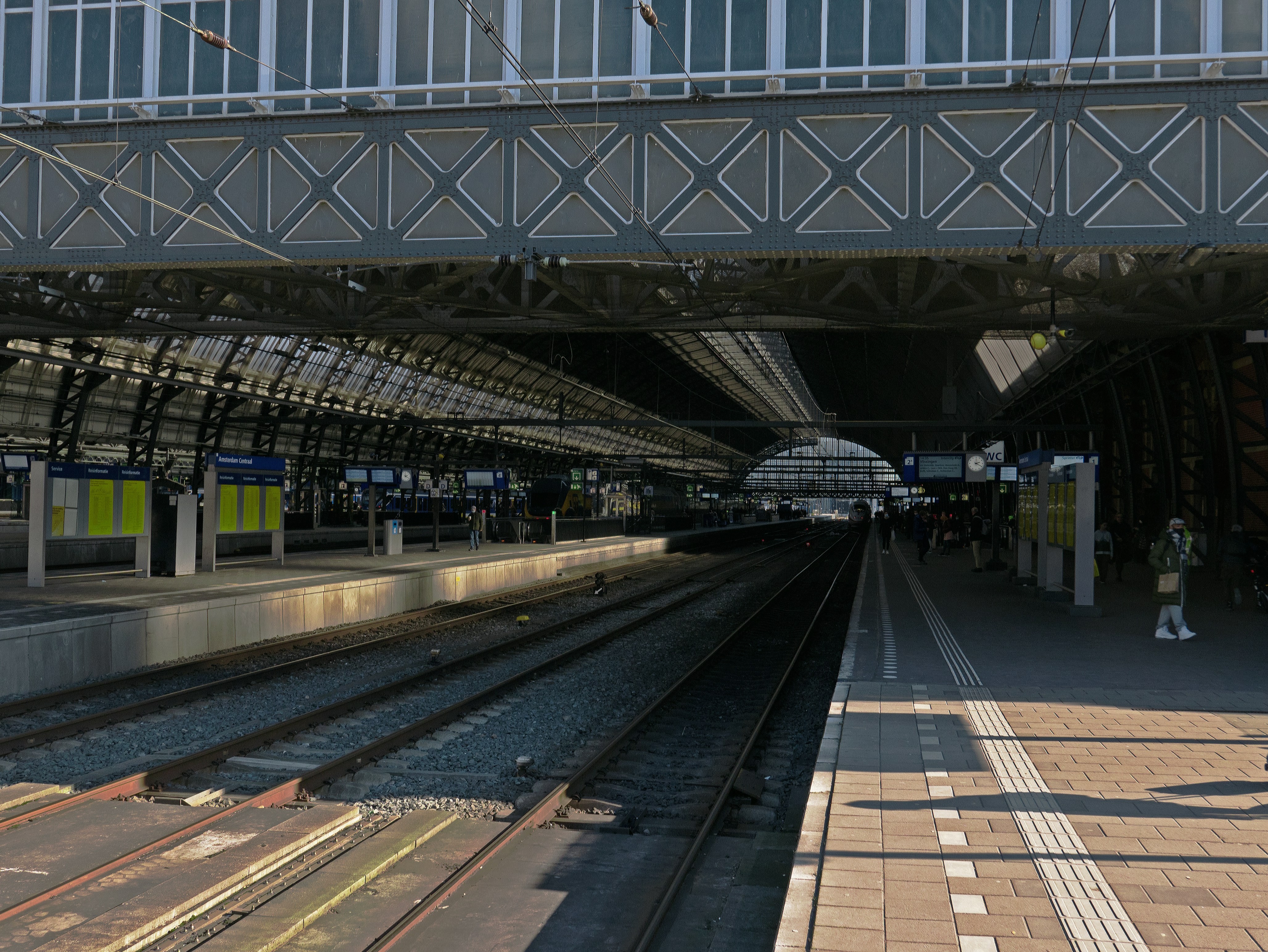 Photo with a view through the long metal cap structure of the old building of Central Station Amsterdam, downtown. In this image I liked to express the long length of the old hall and its pattern of metal beams and windows. This is Dutch historical station architecture of a smoke cap, built c. 1890 for catching the smoke of the steam trains Free station photo of Amsterdam architecture - street photography of The Netherlands of Fons Heijnsbroek March 2022.